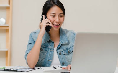 Beautiful smart business Asian woman in smart casual wear working on laptop and talking on phone while sitting on table in creative office. Lifestyle women working at home concept.